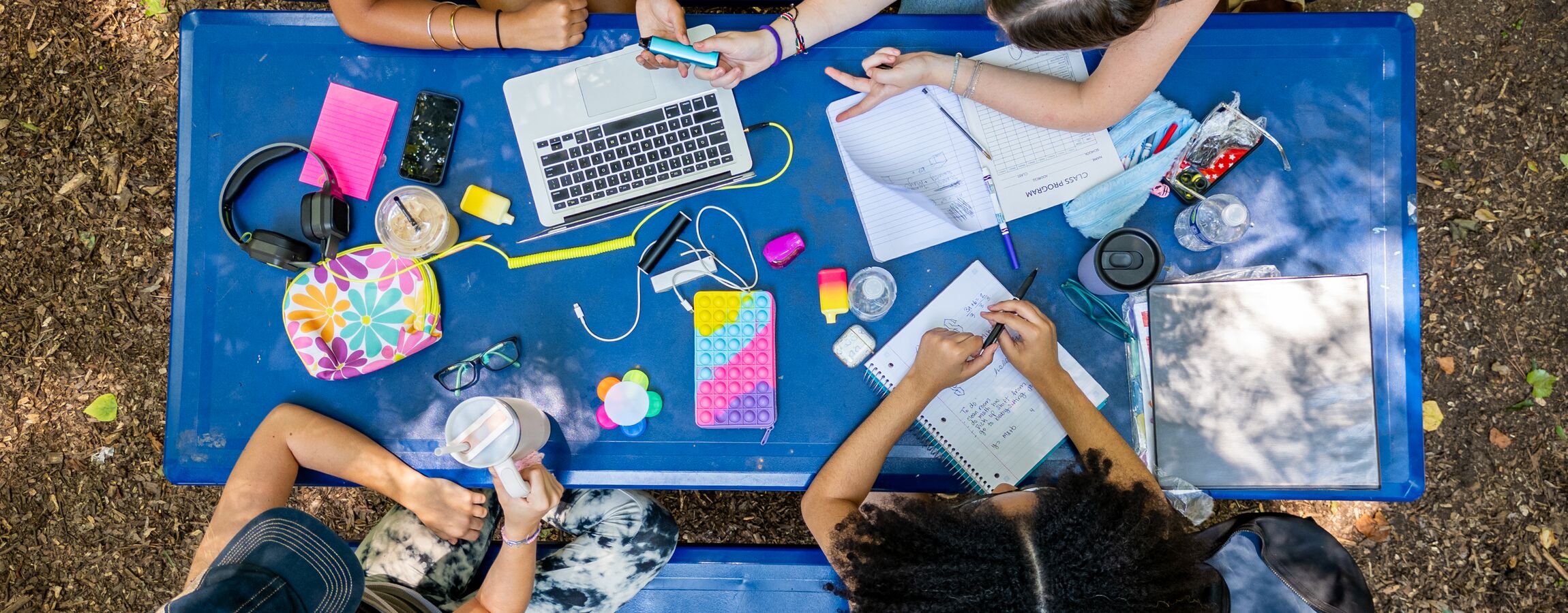 Students studying together outdoors at a blue picnic table, with a laptop, notebooks, phones, headphones, and school supplies spread across the surface, shot from above.